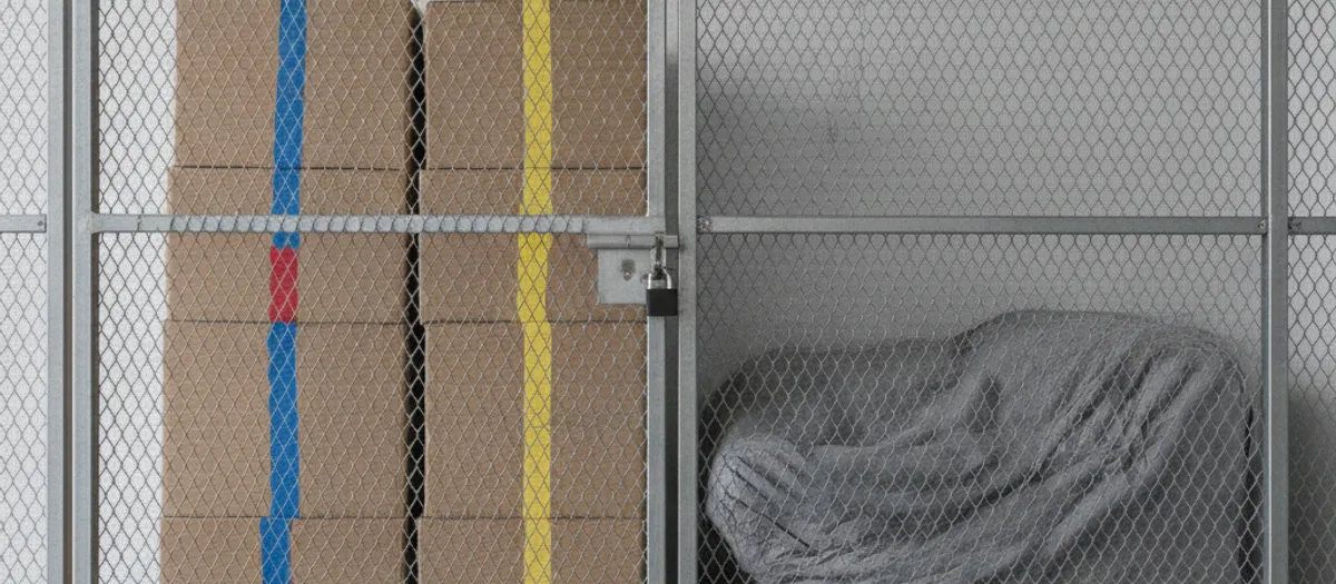 Hinter einer verschlossenen Gittertrennwand stehen gleichmäßig gestapelte Umzugskartons, die nur mit farbigen Klebebändern markiert sind, daneben ein in Decken eingeschlagenes Möbelstück.